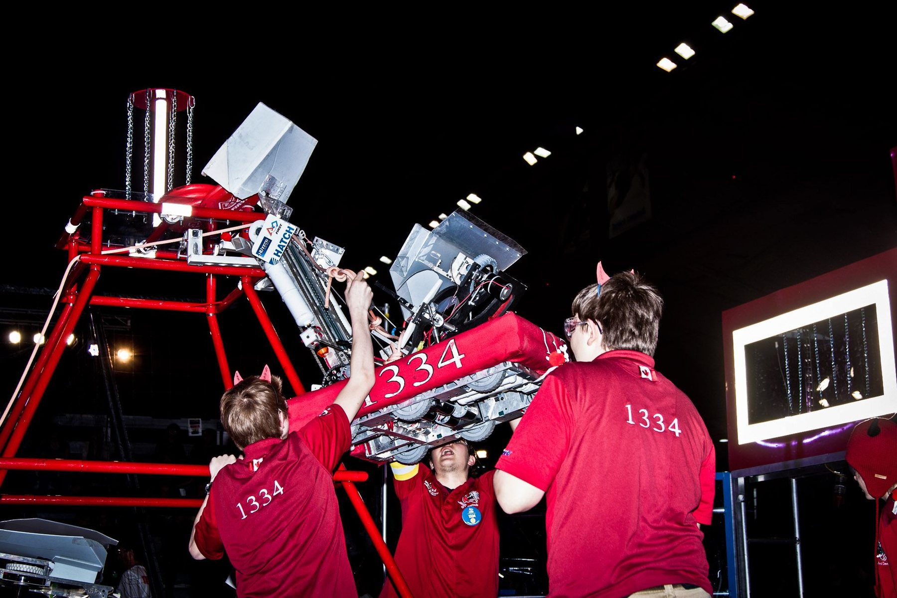 Team 1334 students in red shirts handling their robot against the red competition pyramid