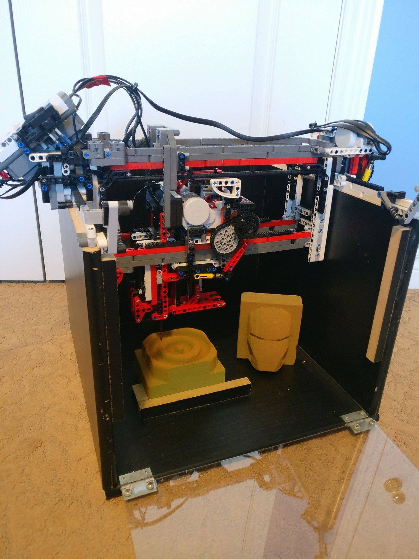The LEGO mill built into an open-fronted black box, its gantry and drill above two carved foam pieces, one an Iron Man helmet