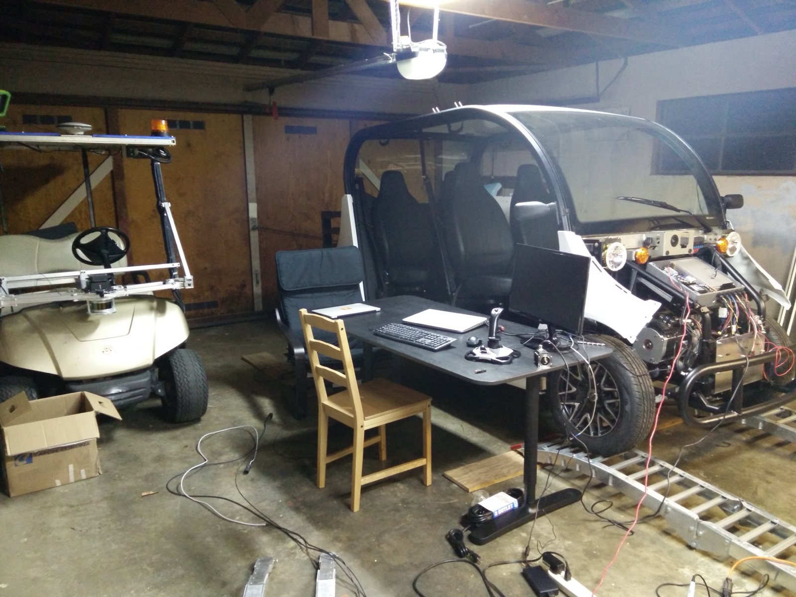 Garage workshop: the Gen 1 golf cart with its roof sensor rack on the left, the Gen 2 Gem on the right with its front panels off and wiring exposed, and a desk with a joystick between them